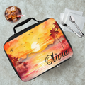 Lunch bag with sunset design and 'Olivia' text on a cafeteria table with a drink and utensils.