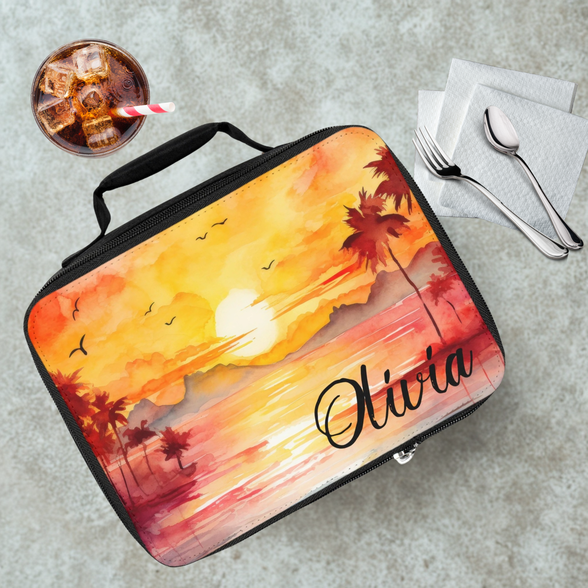 Lunch bag with sunset design and 'Olivia' text on a cafeteria table with a drink and utensils.