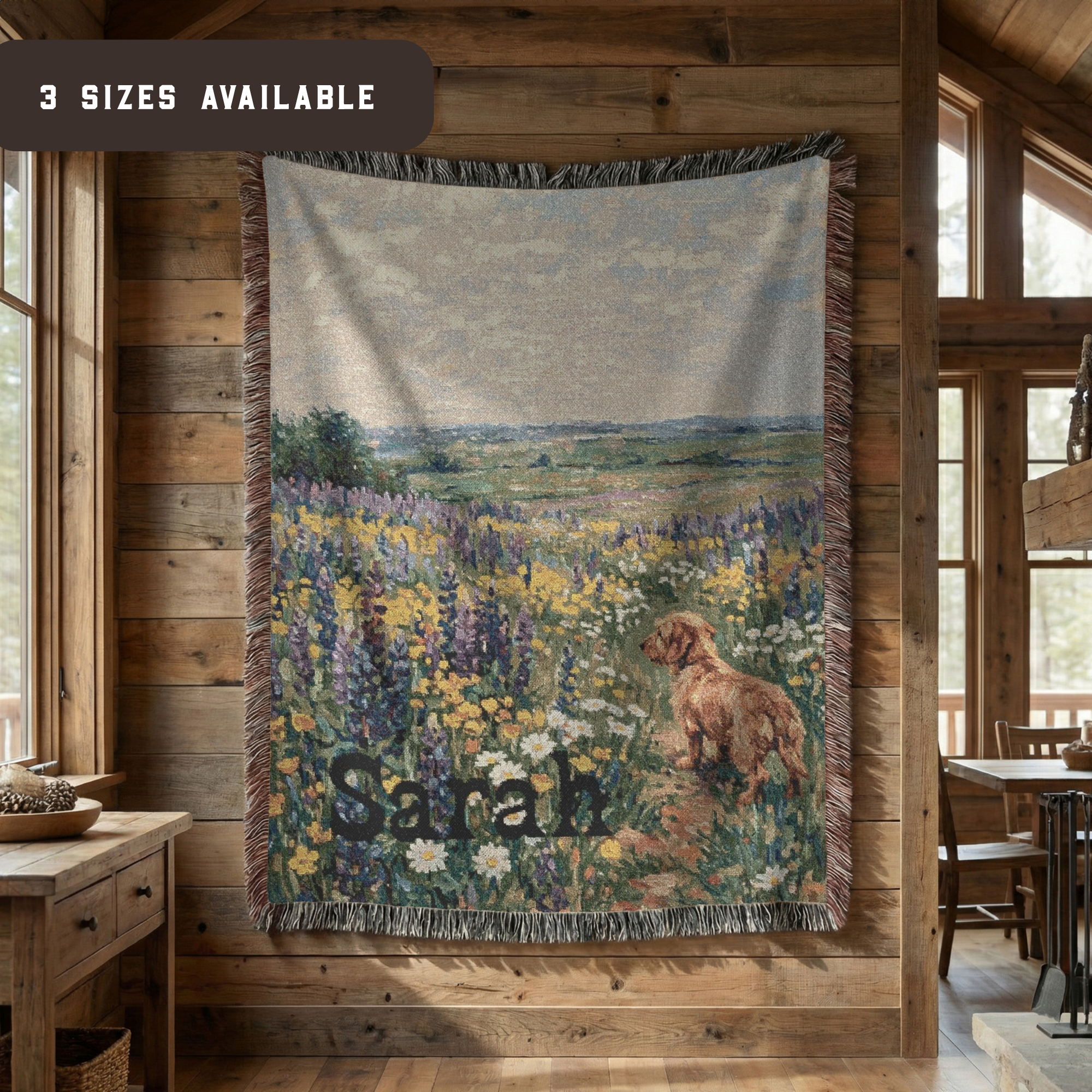 A woven throw blanket with a rustic landscape print of a dog in a flower field, hanging on a wooden wall inside a cabin. The blanket features the name 'Sarah' woven into the design.