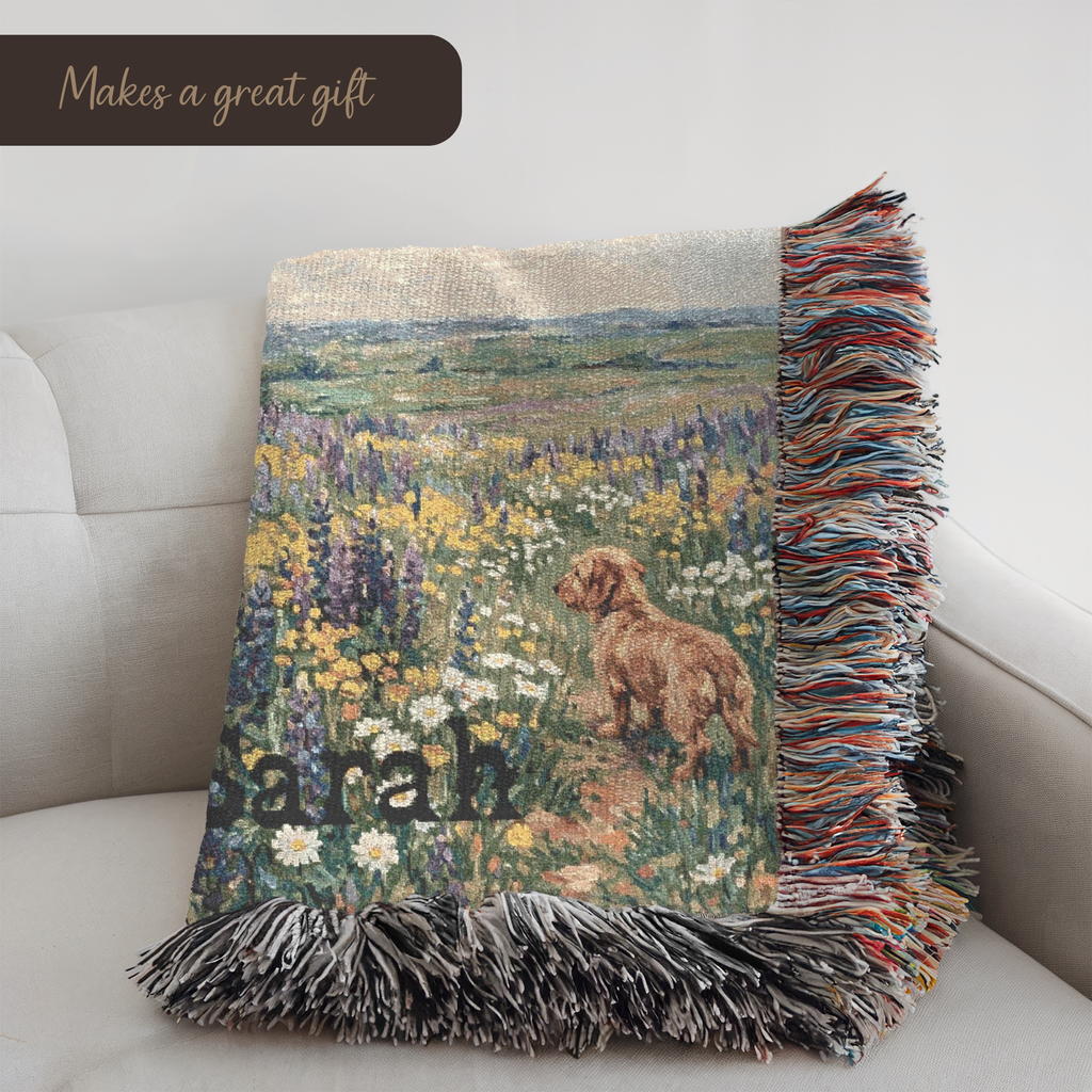 A woven throw blanket with fringe, featuring a printed landscape of a Westie dog in a flower field and the name 'Sarah', styled on a light gray sofa.