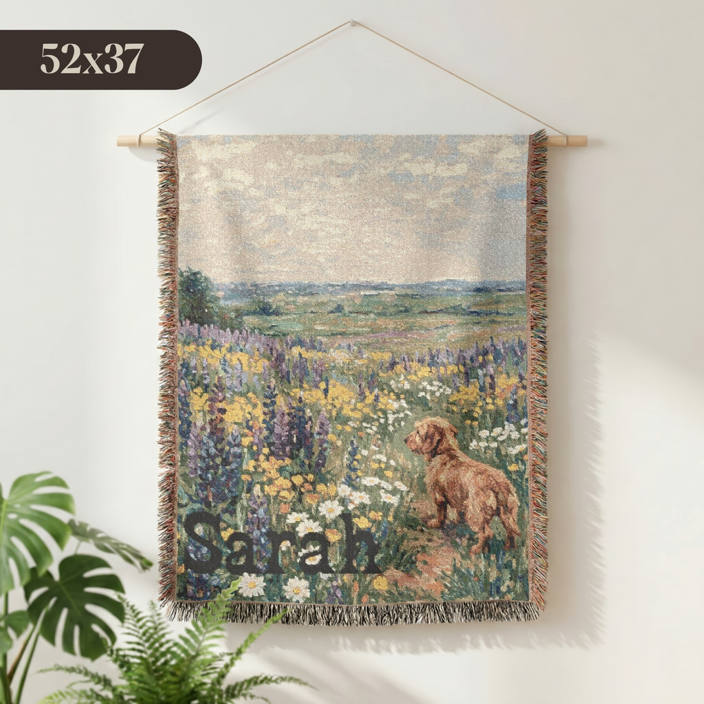 A woven wall tapestry featuring a landscape scene with wildflowers and a dog, hanging on a white wall with a wooden dowel. The tapestry has fringed edges and a dark brown banner at the bottom with the name 'Sarah' in script.