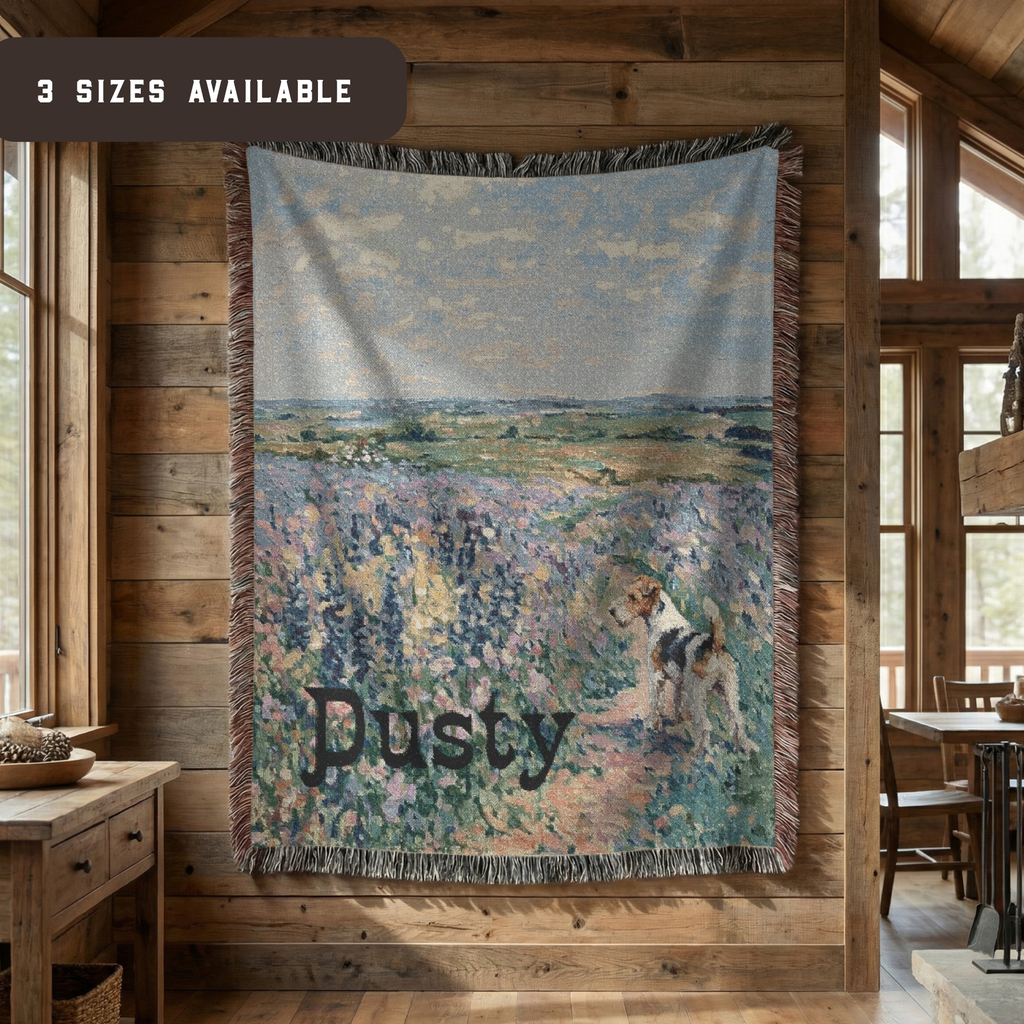A woven throw blanket with fringe is displayed on a rustic wooden wall inside a cabin. The blanket features an impressionistic landscape painting of a dog walking through a field, with the name 'Dusty' printed across the bottom.