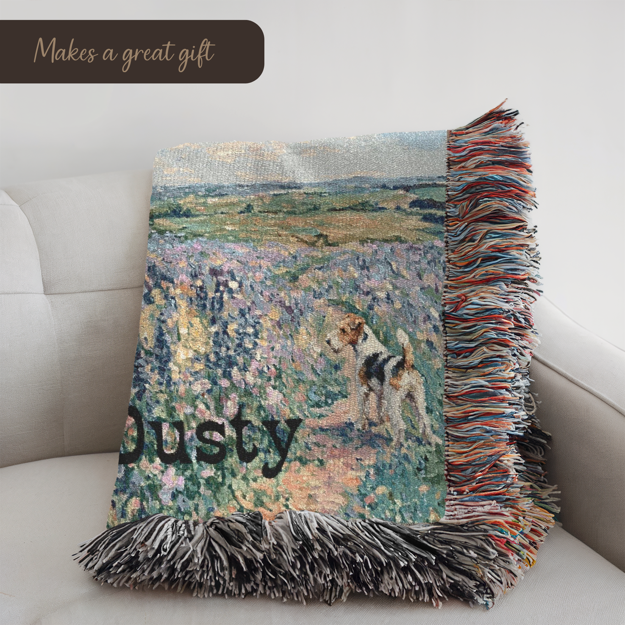 A woven throw blanket draped over a light beige sofa, featuring an impressionistic landscape print with a dog and the word 'Dusty' in black. The blanket has multicolored fringe on the right and bottom edges, and a dark brown banner in the top left corner reads 'Makes a great gift'.
