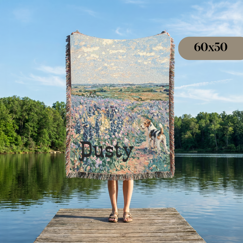 A large woven throw blanket featuring an impressionistic landscape of a dog in a flower field is held up by a person standing on a wooden dock by a lake. The blanket has fringe on the sides and the word 'Dusty' printed across the lower portion.