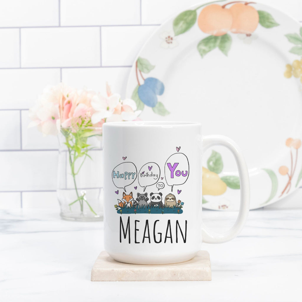 White mug with 'Happy Birthday You Meagan' text and animal illustrations on a white surface with a decorative plate in the background.