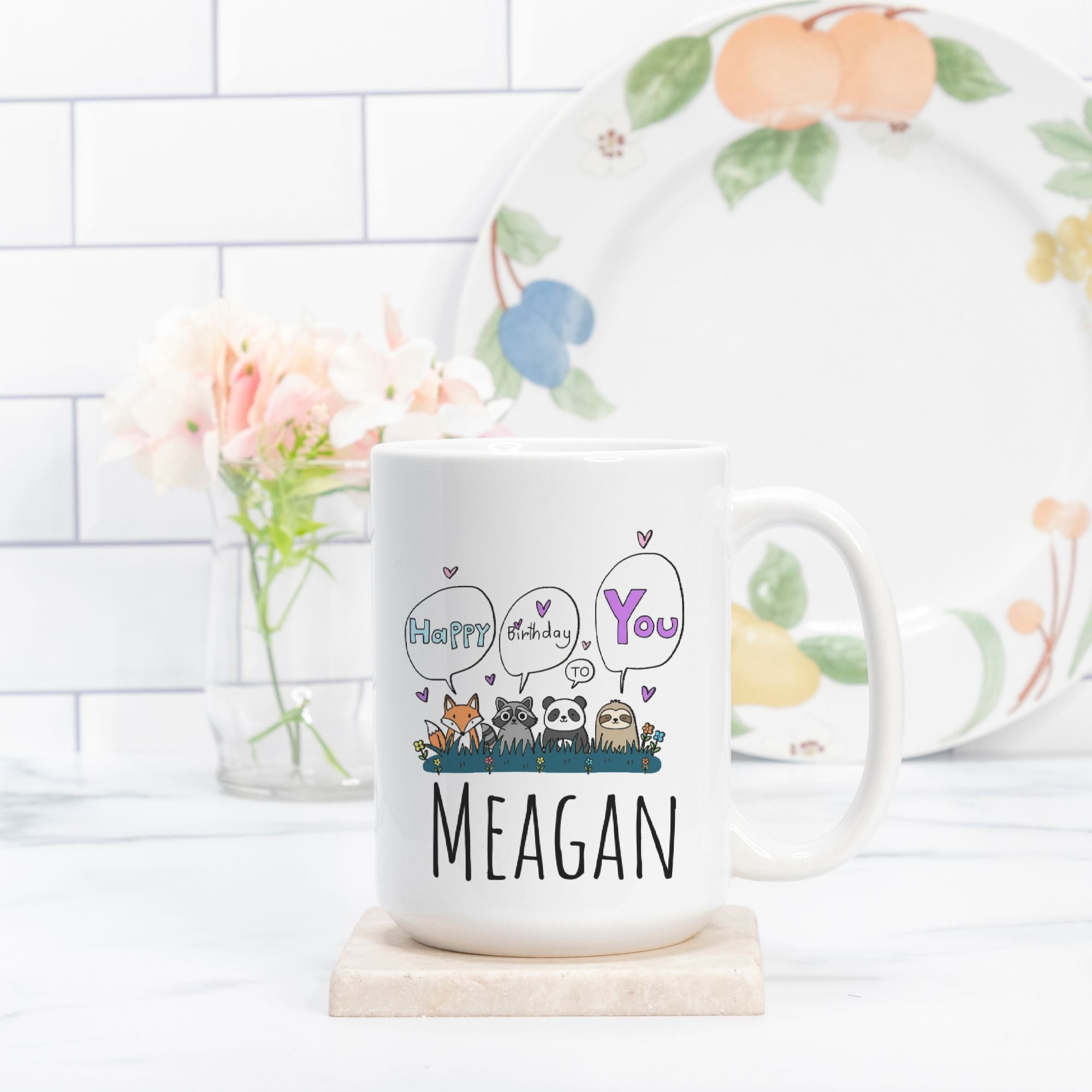 White mug with 'Happy Birthday You Meagan' text and animal illustrations on a white surface with a decorative plate in the background.