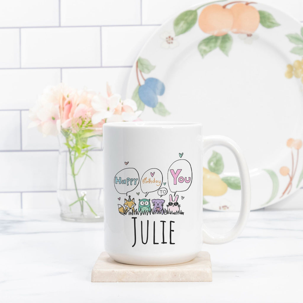 White mug with 'Happy Birthday You' and 'Julie' text, placed on a wooden coaster with a floral plate in the background.