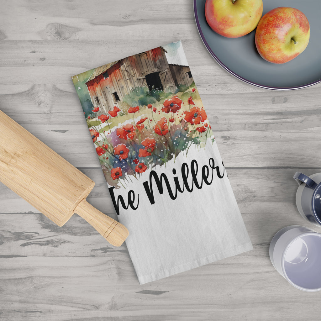 Tea towel with floral barn design and 'The Miller' text on a wooden surface with apples and a rolling pin.