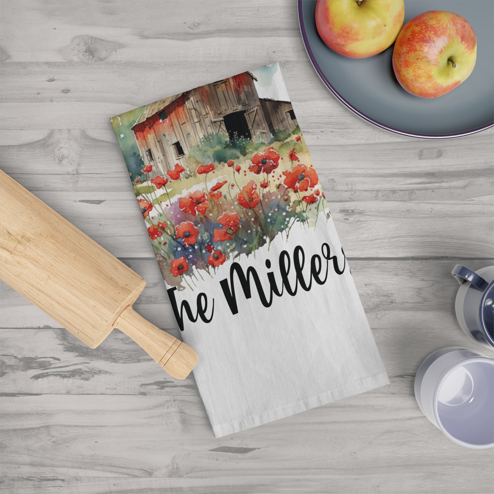 Tea towel with floral barn design and 'The Miller' text on a wooden surface with apples and a rolling pin.
