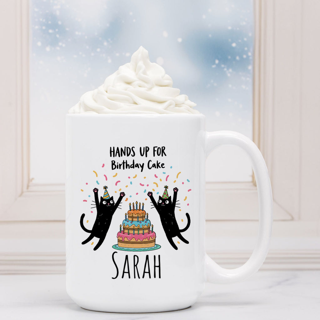 White mug with black cats and birthday cake design, text 'Hands up for Birthday Cake Sarah', against a blurred background of whipped cream and a blue sky.