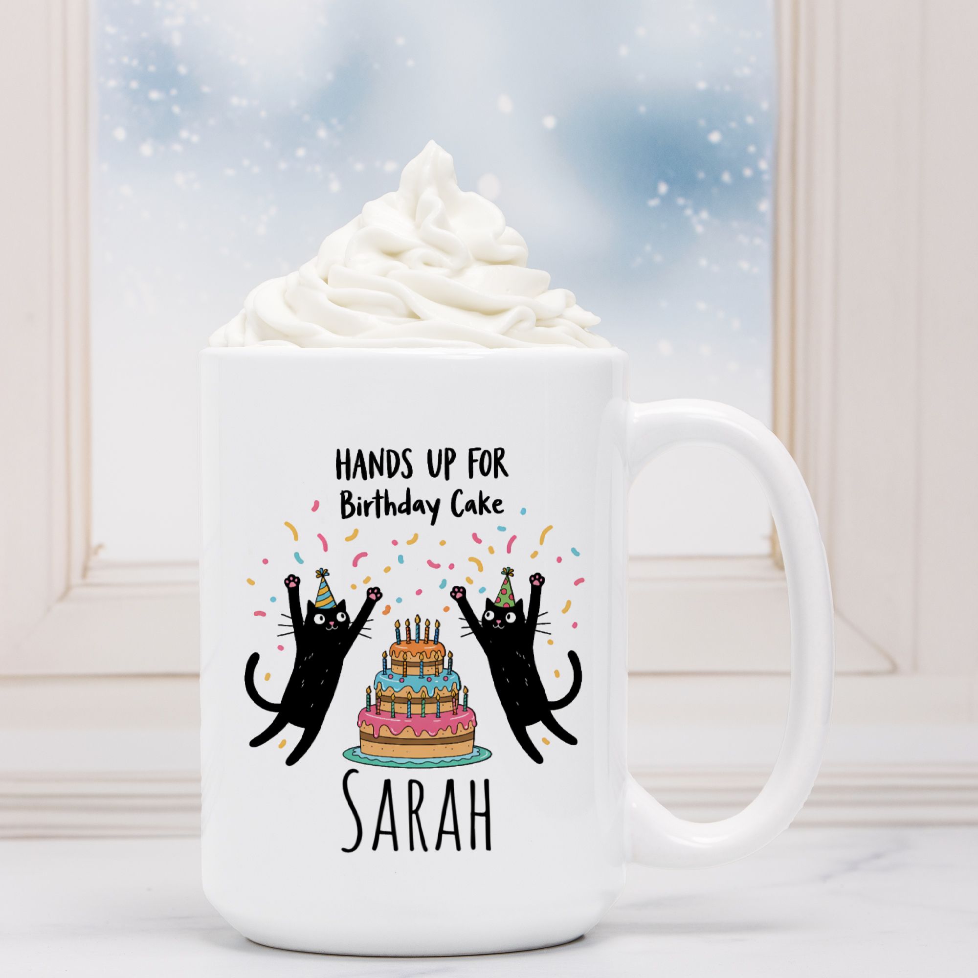 White mug with black cats and birthday cake design, text 'Hands up for Birthday Cake Sarah', against a blurred background of whipped cream and a blue sky.