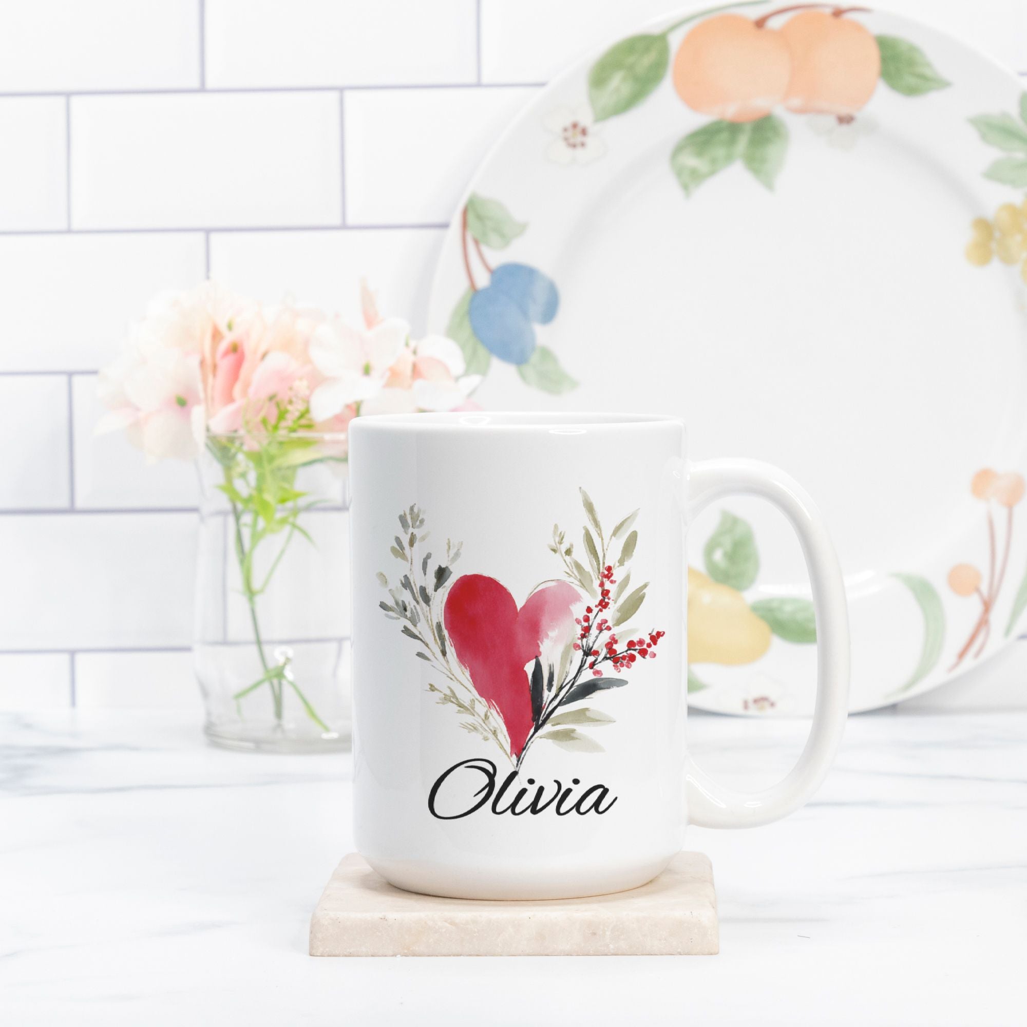 White mug with floral design and heart, featuring the name 'Olivia', on a white surface with a decorative plate in the background.
