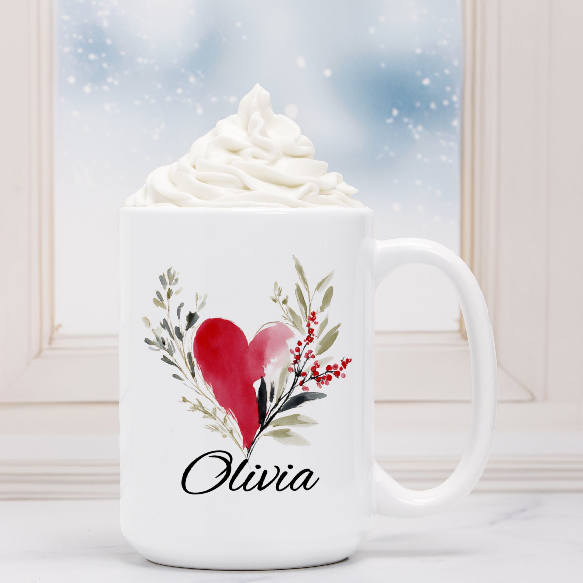 White mug with a red heart and green leaves design, filled with hot chocolate and whipped cream.