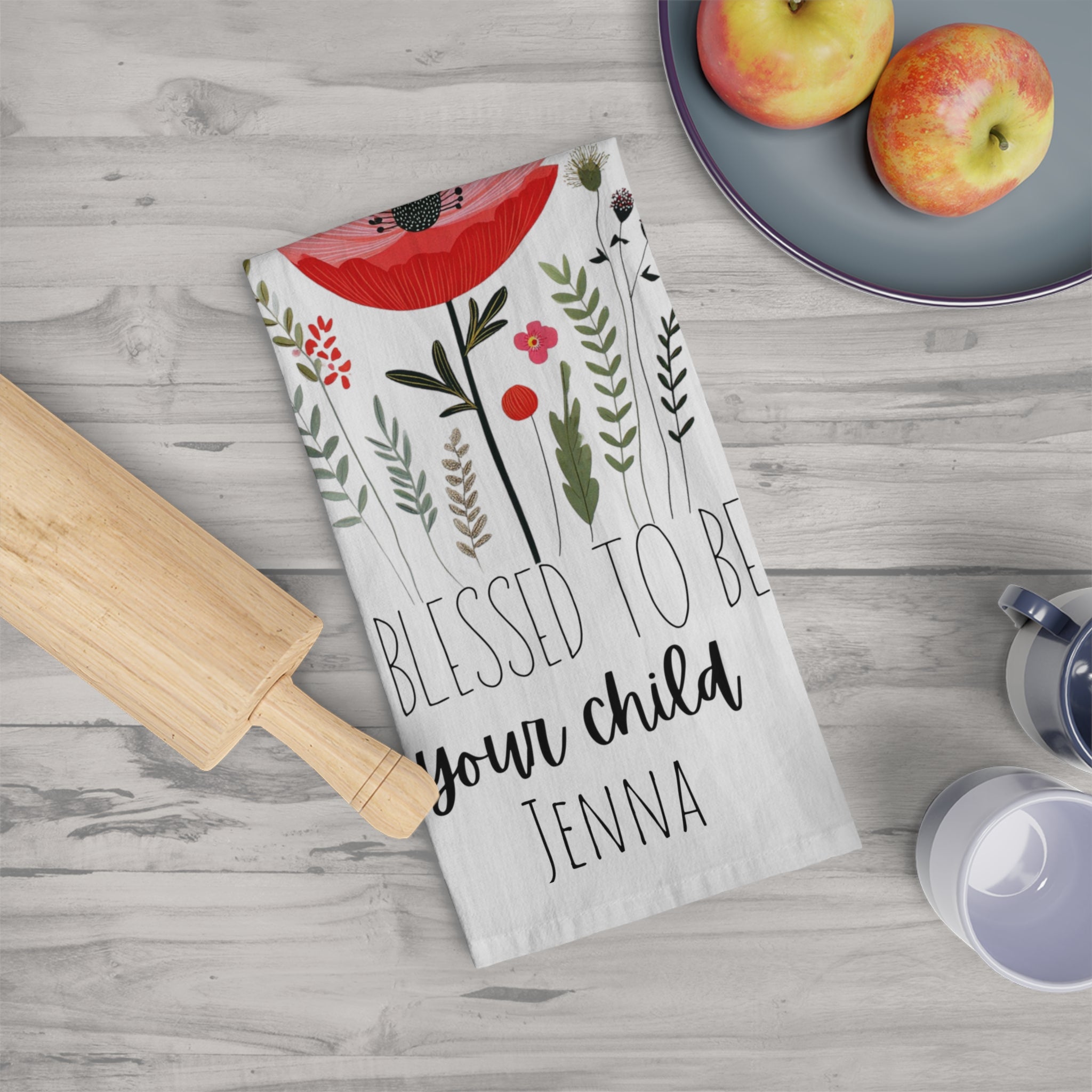 Kitchen towel with floral design and text on a wooden surface with apples and a rolling pin.