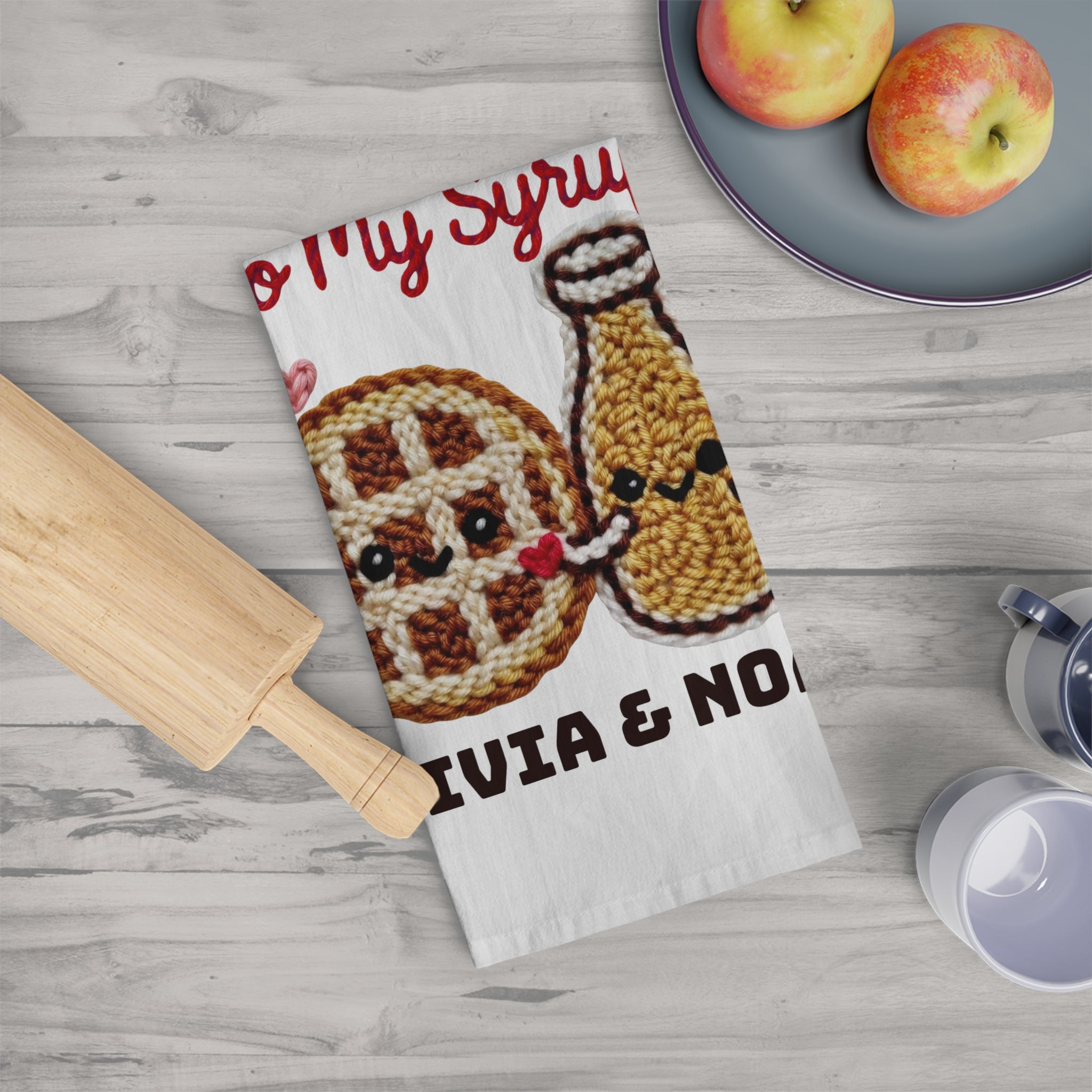 Tea towel with waffle and syrup design on a wooden surface with apples and a rolling pin.
