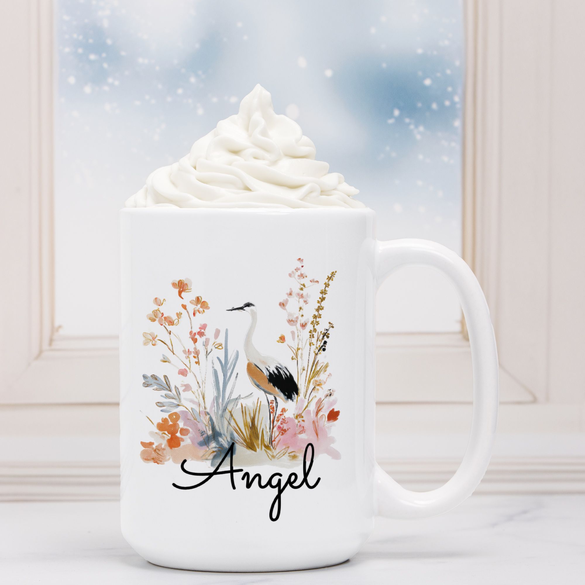White mug with a bird and floral design labeled 'Angel' against a snowy background.