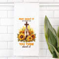 Decorative tea towel with cross, praying hands, and sunflowers on a white tiled wall.