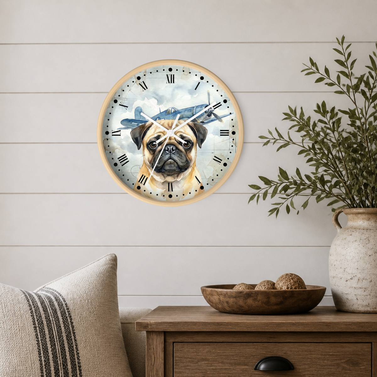 A round wall clock with a light wood frame hangs on a white shiplap wall above a wooden side table. The clock face features a watercolor illustration of a pug dog and a blue vintage airplane against a cloudy sky.