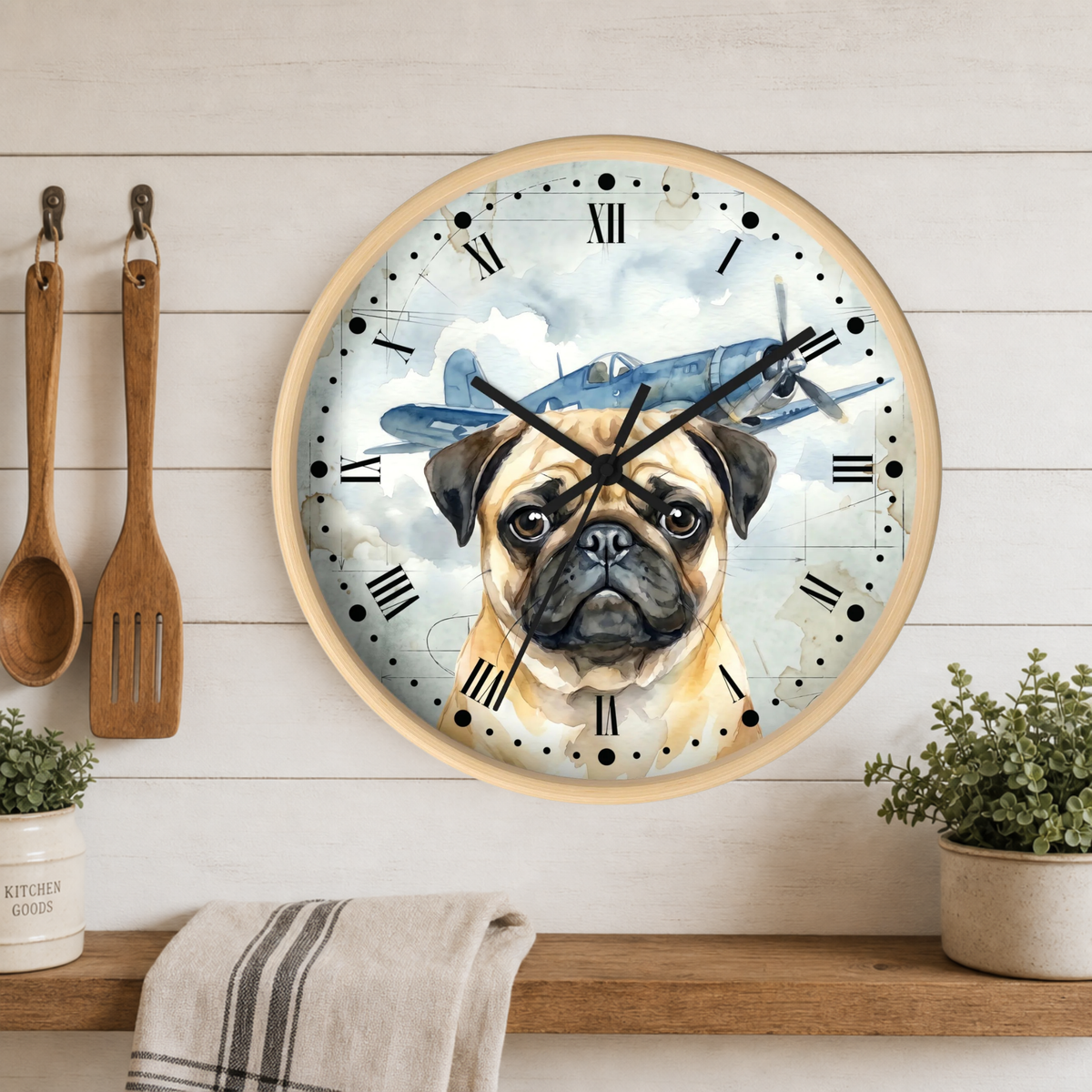 Round wall clock with a light wood frame, featuring a printed design of a pug dog and a vintage airplane on the face. The clock is mounted on a white wood-paneled wall above a wooden shelf with kitchen utensils, a towel, and small potted plants.