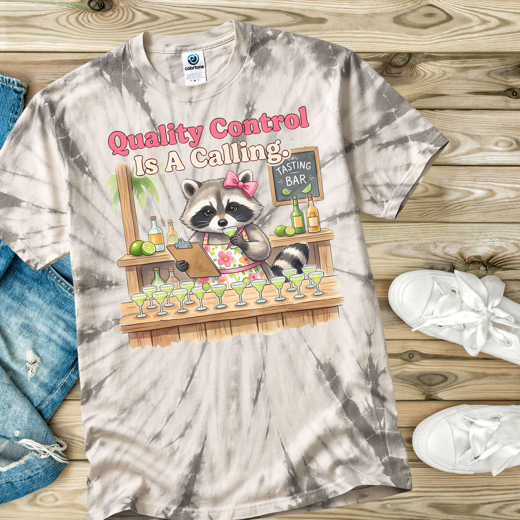 Tie-dye t-shirt with a raccoon character and 'Quality Control is a Calling' text on a wooden surface.