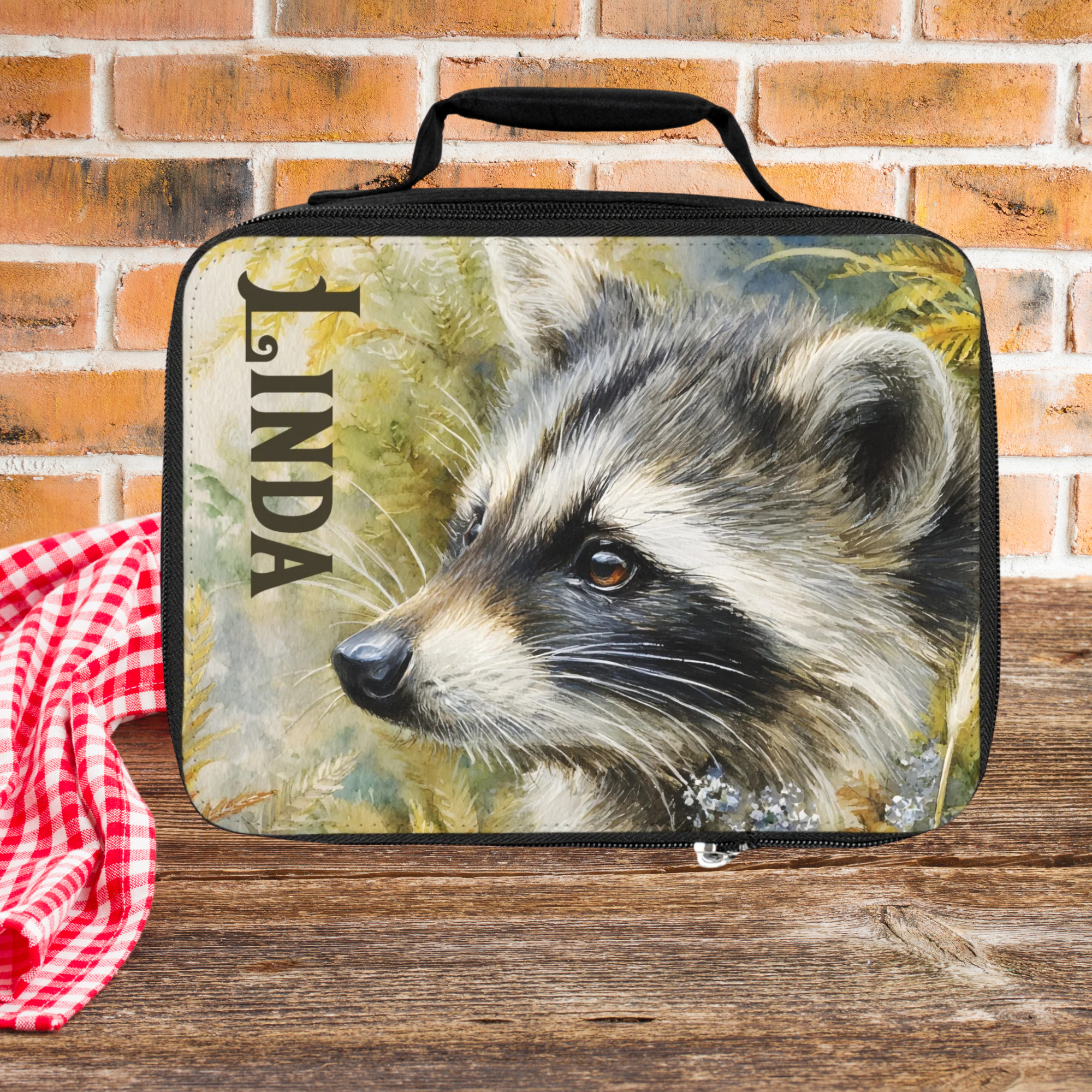 Lunch bag with raccoon design and 'LINDA' custom text on a wooden picnic table against a  brick background