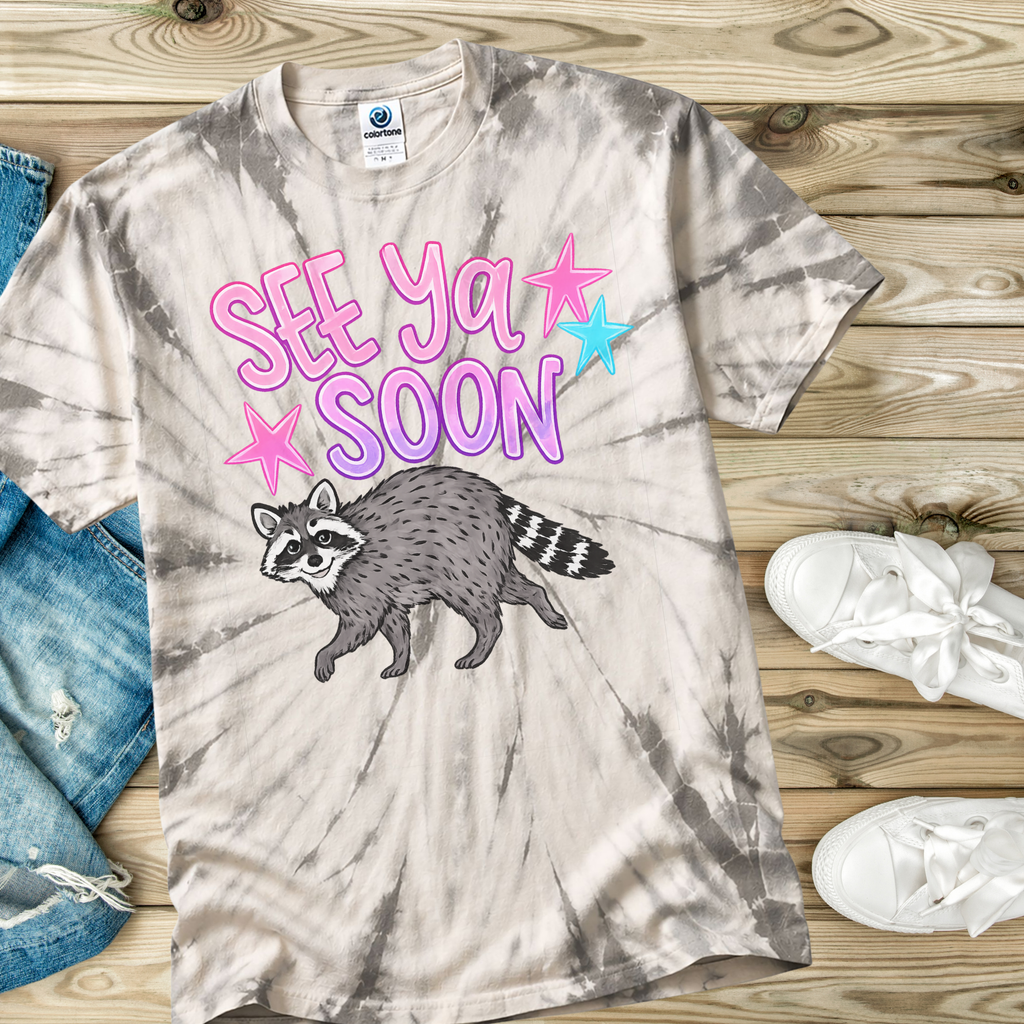 Spider Silver Tie-dye t-shirt with raccoon graphic and 'See Ya Soon' text on a wooden surface.