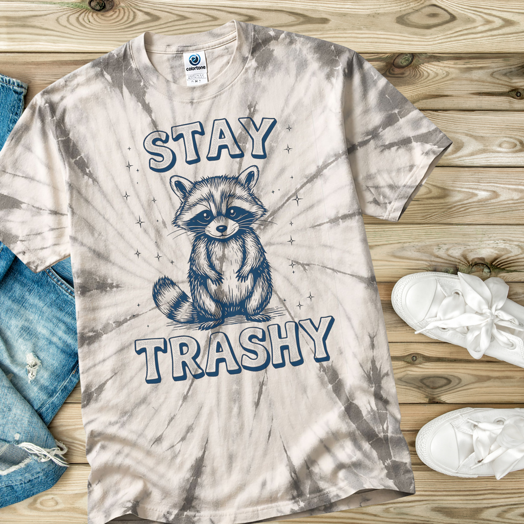 Tie-dye t-shirt with raccoon graphic and 'Stay Trashy' text on a wooden surface.