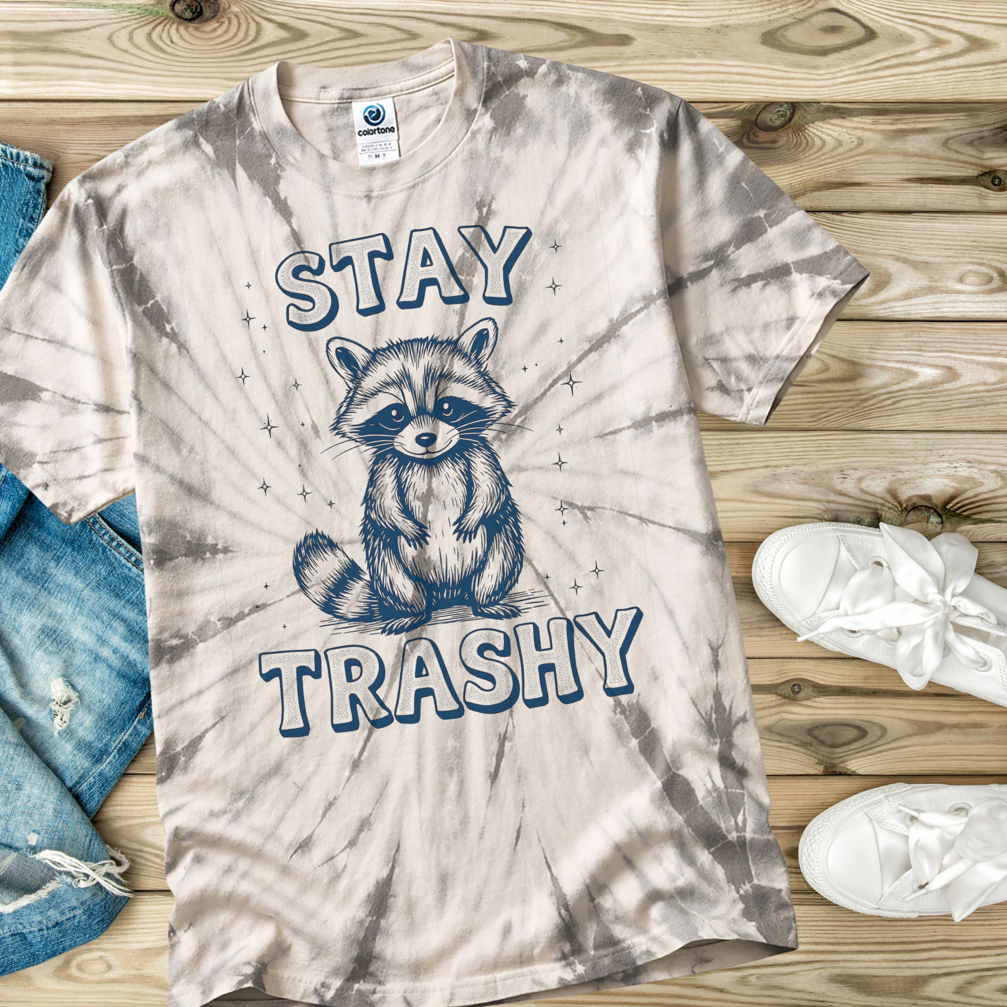 Tie-dye t-shirt with raccoon graphic and 'Stay Trashy' text on a wooden surface.