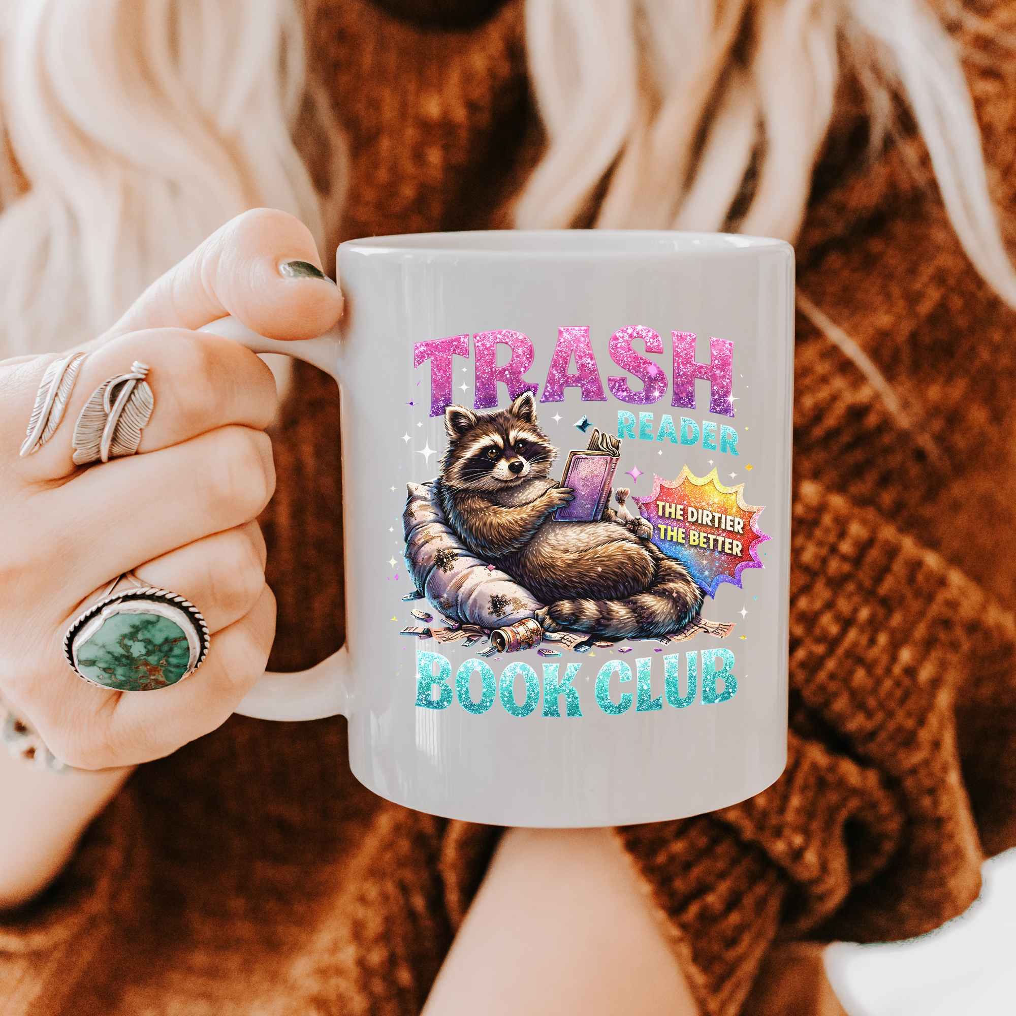 Person holding a mug with a raccoon graphic and text 'Trash Reader Book Club' against a blurred background.