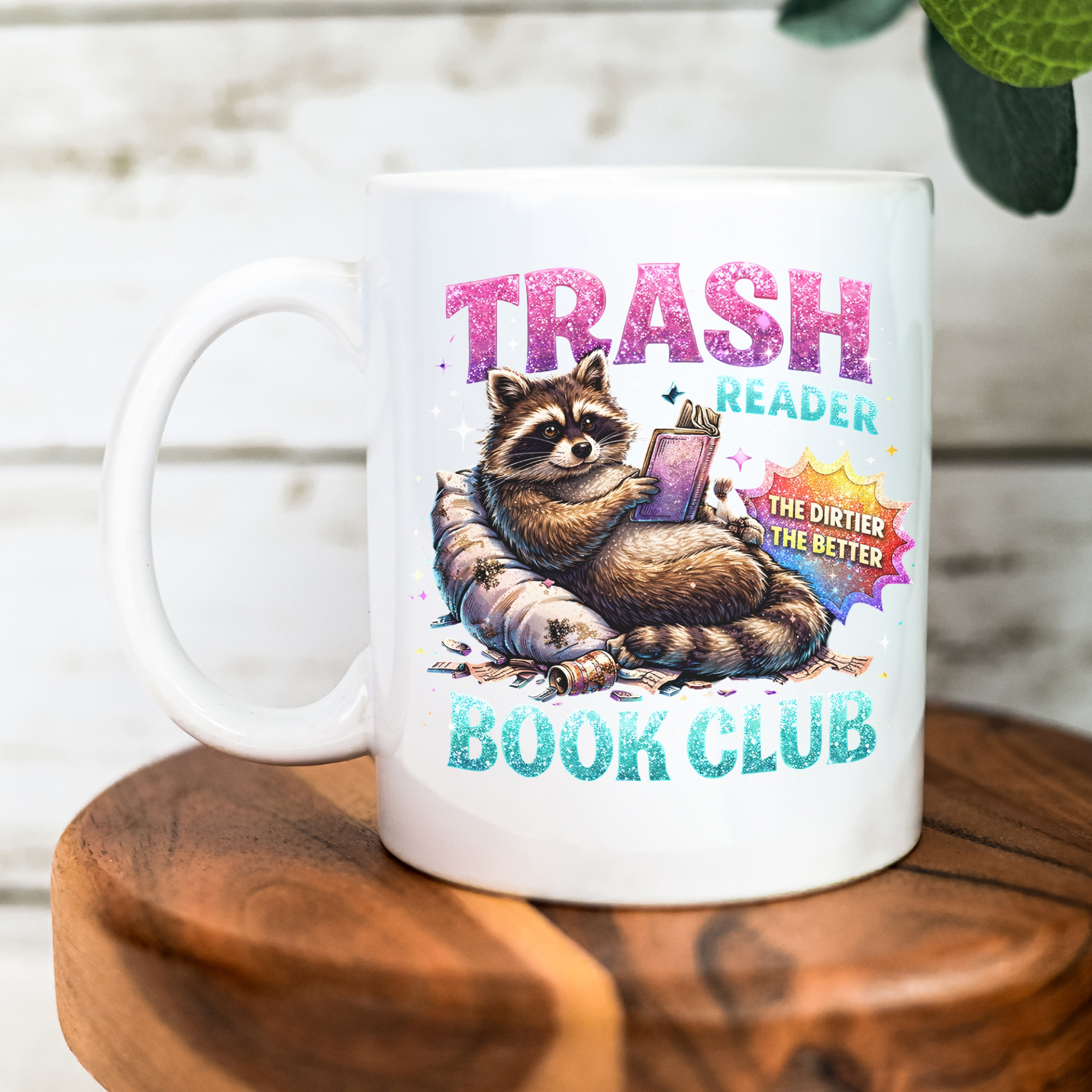 White mug with a raccoon graphic and text 'Trash Reader Book Club' on a wooden surface.