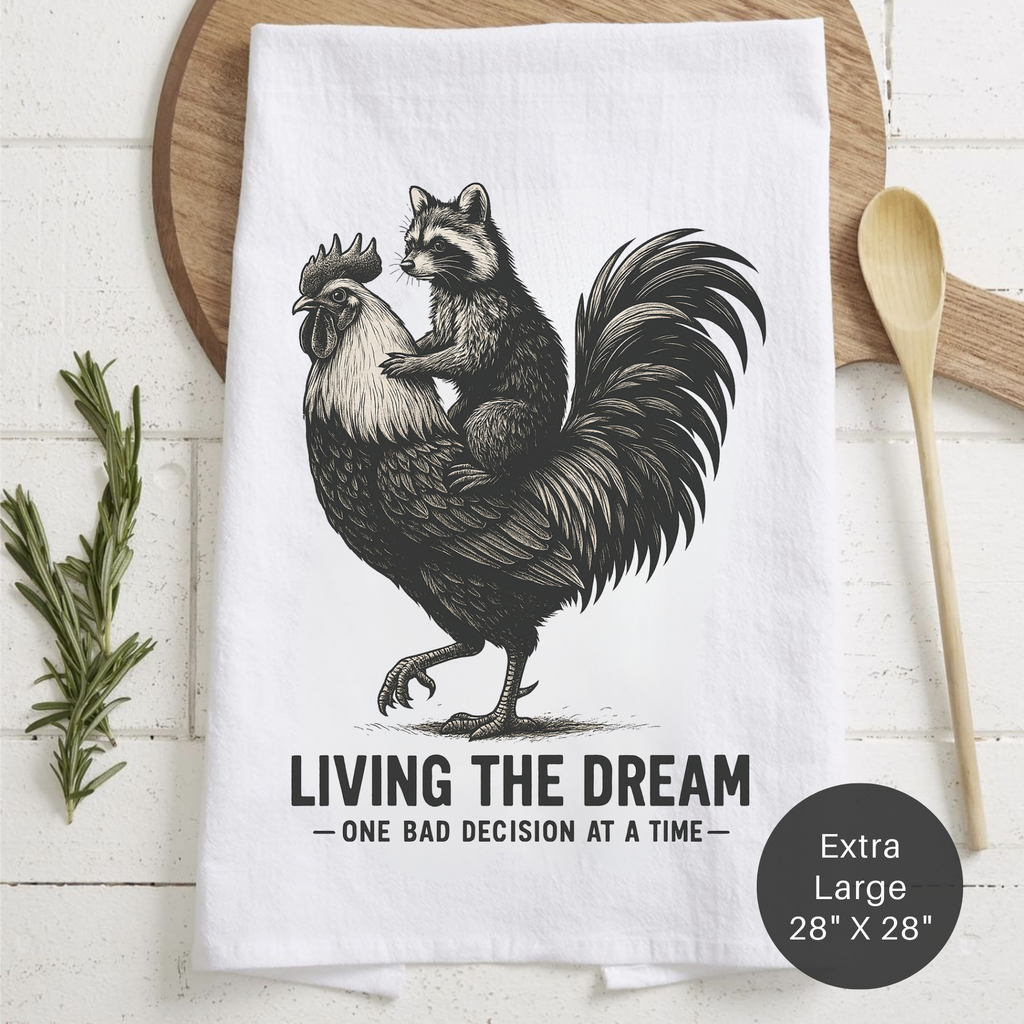 Tea towel with raccoon on rooster design and text 'Living the Dream' on a wooden surface.