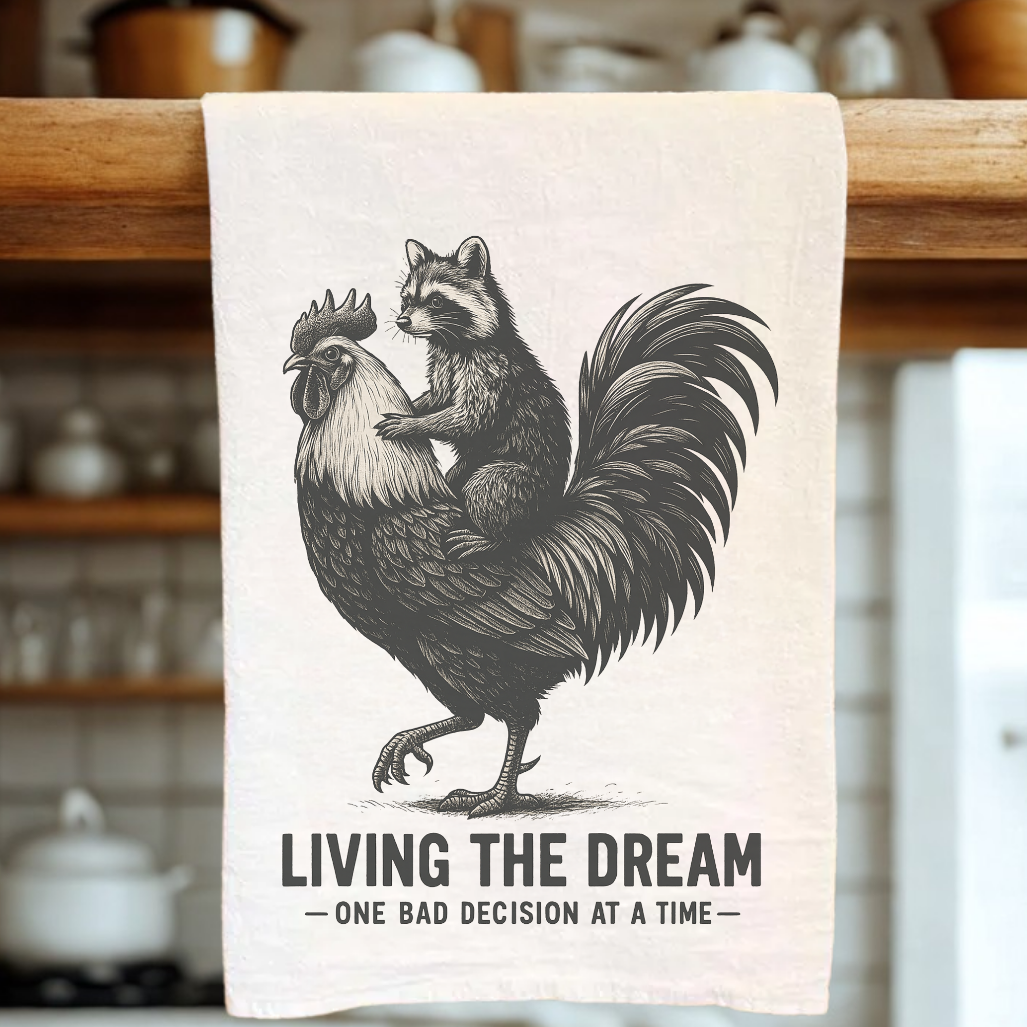 Tea towel with a graphic of a raccoon on a rooster with text 'Living the Dream, One Bad Decision at a Time' in a kitchen setting.