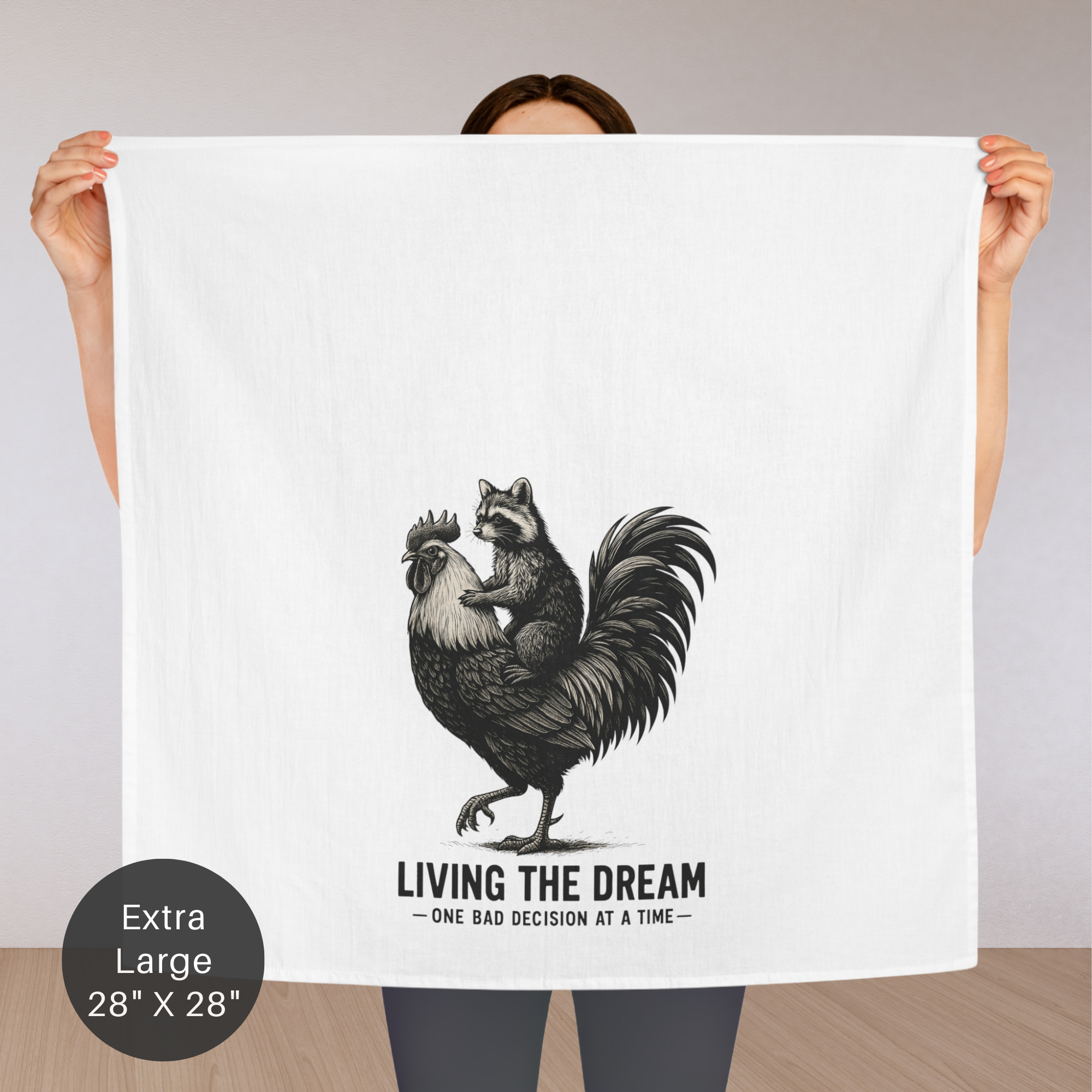 Person holding an extra large tea towel with a graphic of a raccoon riding a rooster, text 'Living the Dream' and 'One bad decision at a time', on a plain background.