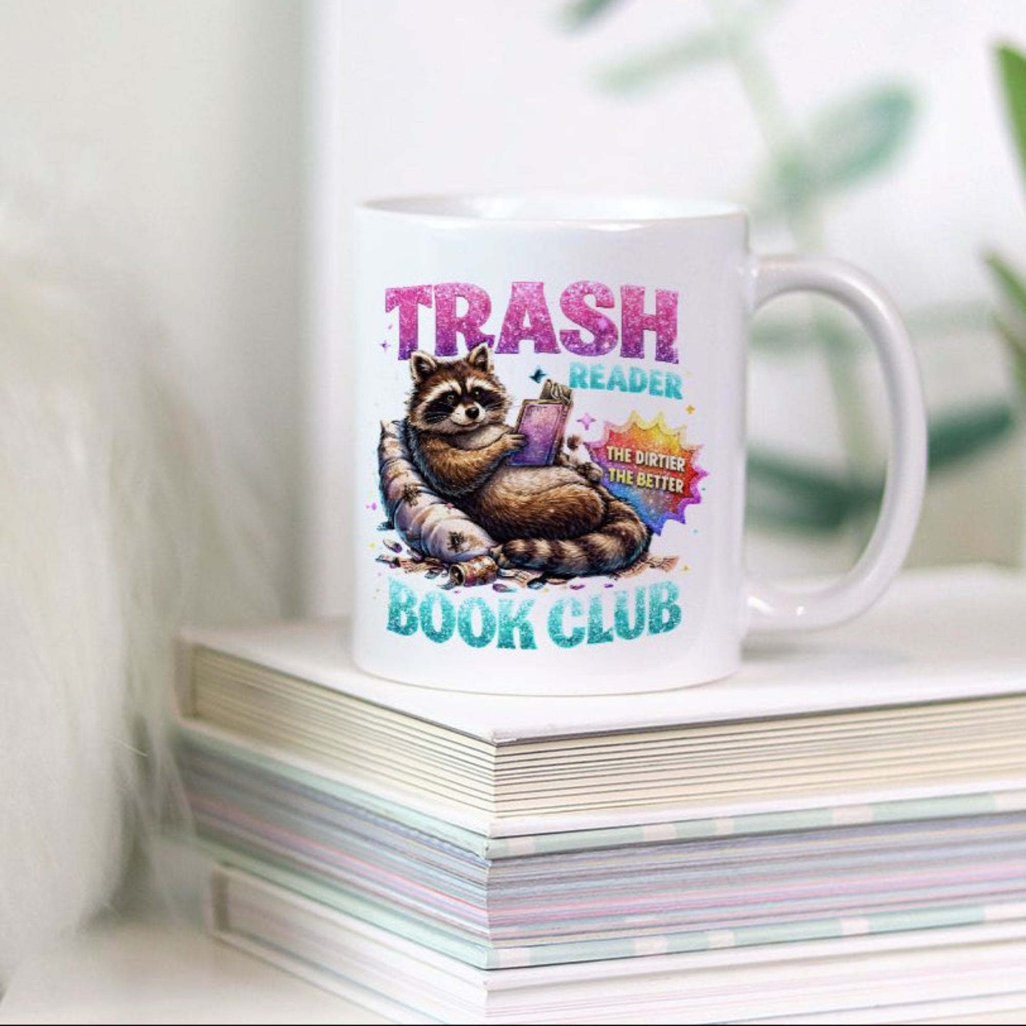 Mug with raccoon graphic and text 'Trash Reader Book Club' on a stack of books with a plant in the background.