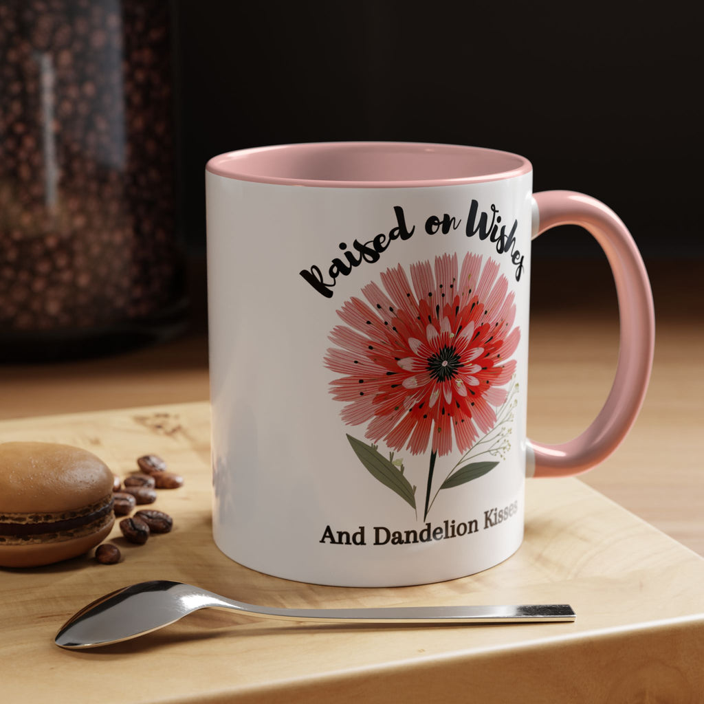 White mug with pink interior and handle, featuring a red flower design and text, on a wooden surface with coffee beans and a spoon.