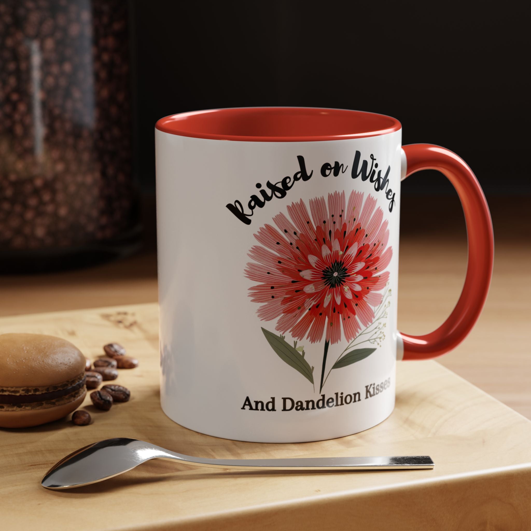 White mug with red interior and handle, featuring a pink flower design and text, on a wooden surface with coffee beans and a spoon.