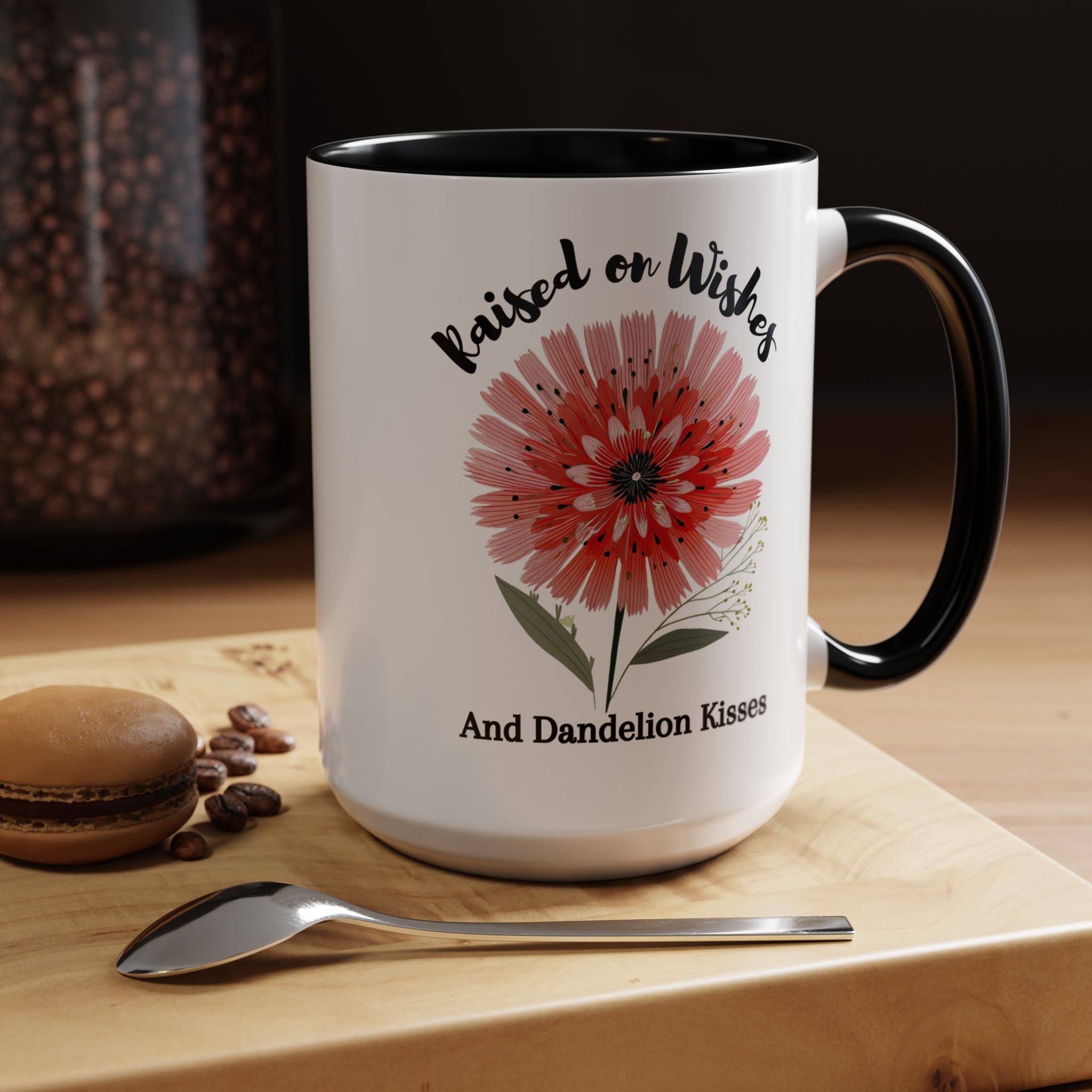 White mug with black interior and handle, featuring a red flower design and text 'Raised on Wishes and Dandelion Kisses', on a wooden surface with coffee beans and a spoon.