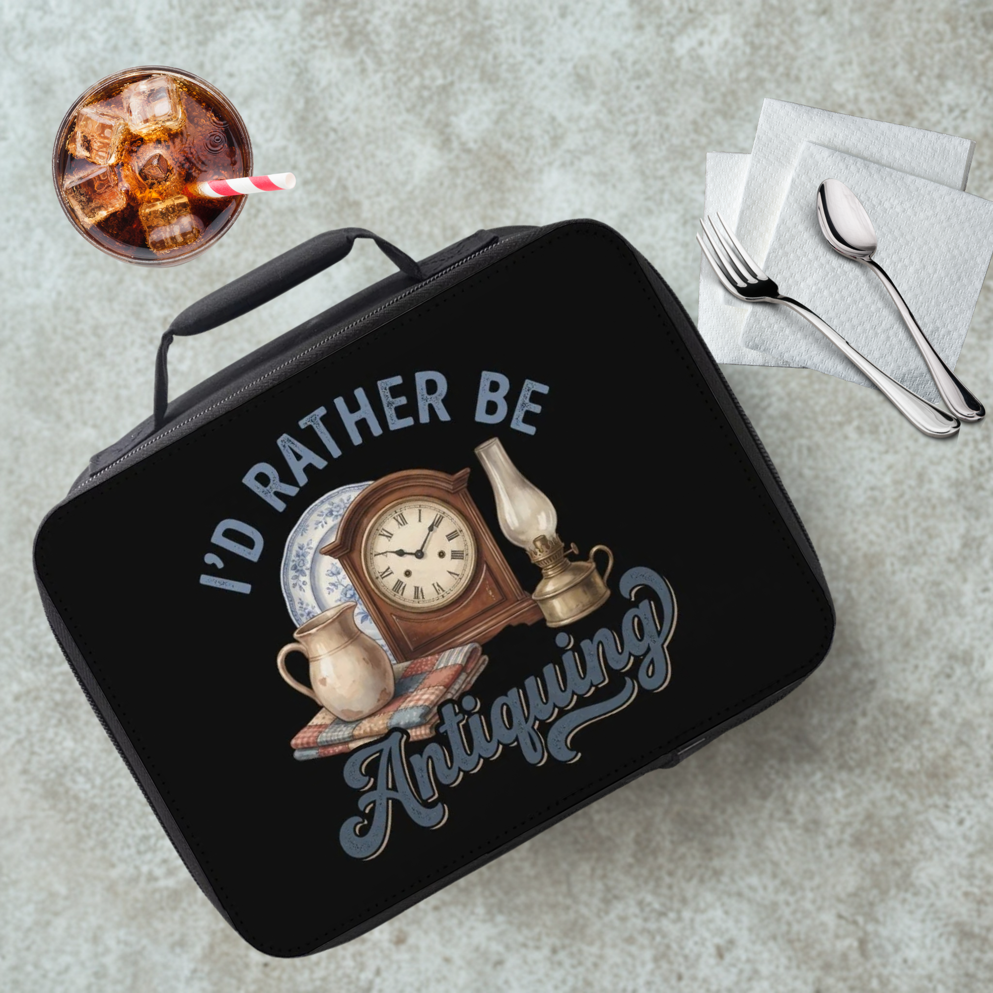 Black lunch box with 'I'd Rather Be Antiquing' design on a gray table with a drink and utensils.