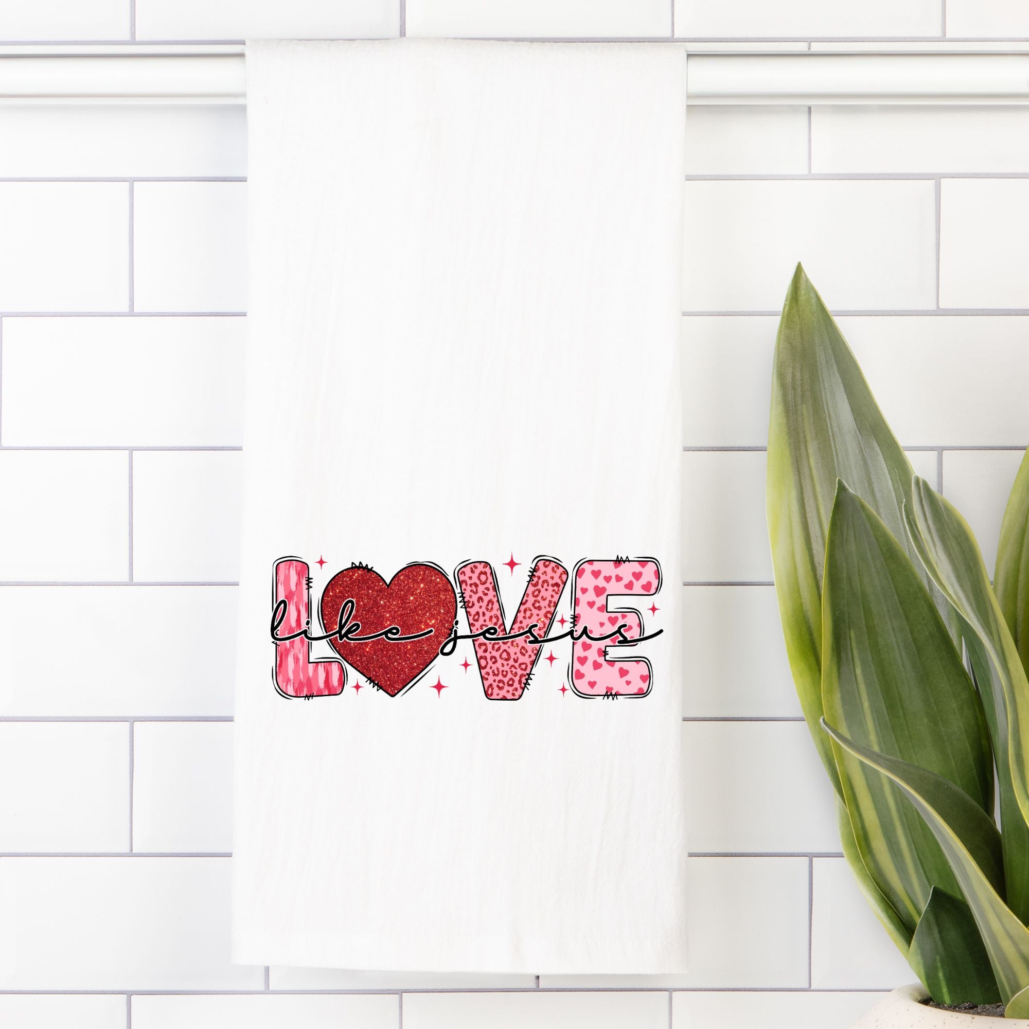 White tea towel with decorative text 'Love Like Jesus' with a heart on a white tiled wall.
