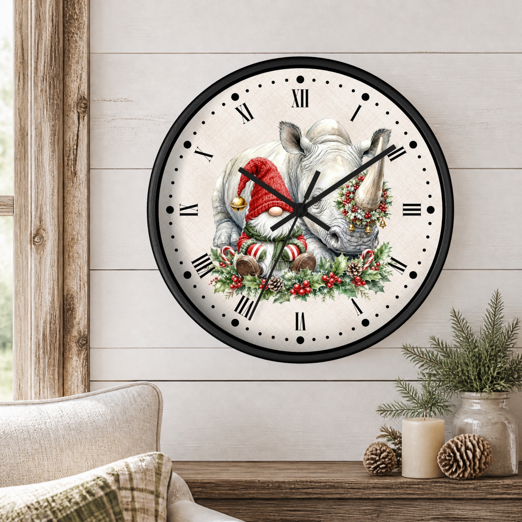A round wall clock with a black frame and a white face, featuring a rhinoceros wearing a Santa hat and holiday wreath. The clock is styled in a cozy room setting on a light wood-paneled wall above a wooden table with pine branches and candles.