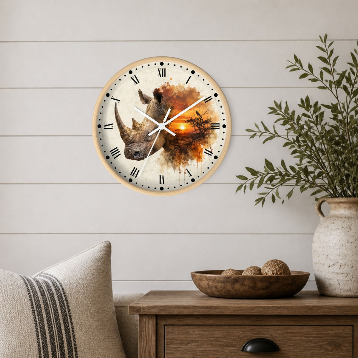 A round wall clock with a rhinoceros and orange watercolor design, mounted on a white shiplap wall above a wooden side table with a plant and decorative bowl.