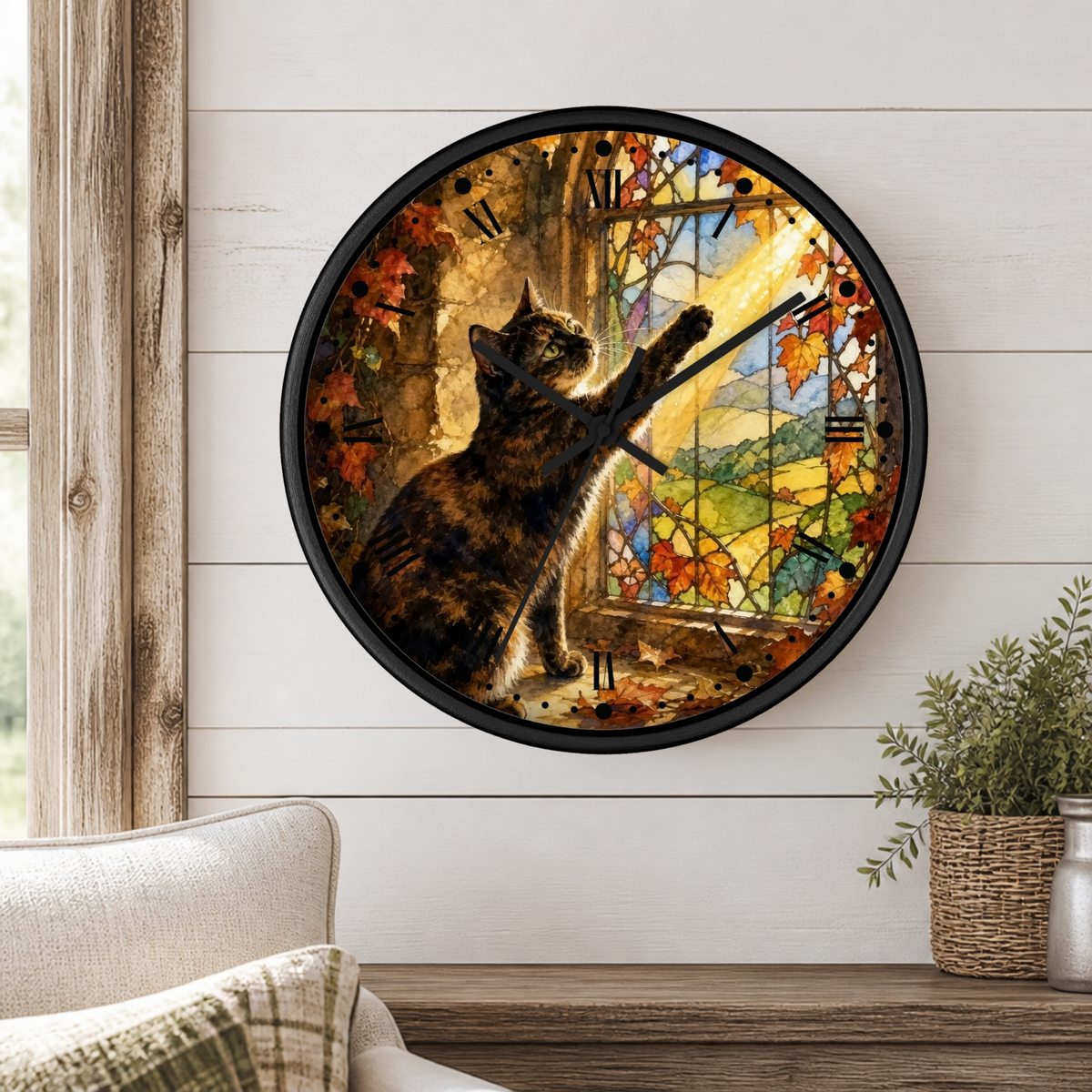 A round wall clock with a black frame and a printed illustration of a tortoiseshell cat stretching its paw toward a colorful autumn landscape window. The clock is mounted on a white shiplap wall above a wooden mantel, with a beige armchair and a potted plant visible in the room.