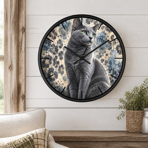 Round wall clock with a black frame featuring a printed illustration of a gray cat over a blue and beige watercolor background, mounted on a white shiplap wall above a wooden console table with a plant and chair.