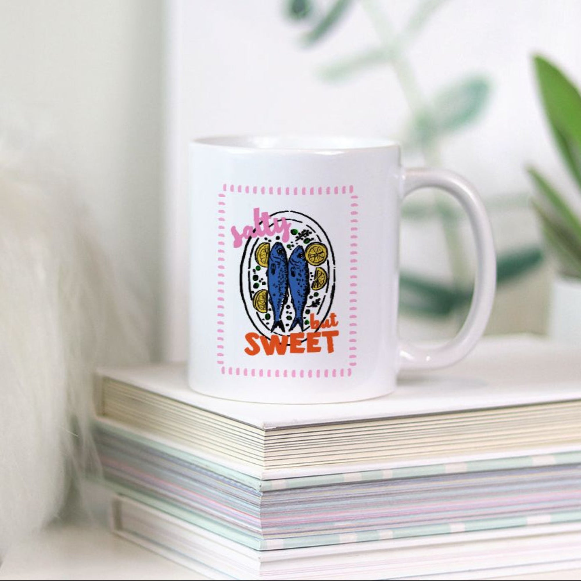 White 11oz. mug with colorful summer sardine design on a stack of books with a plant in the background