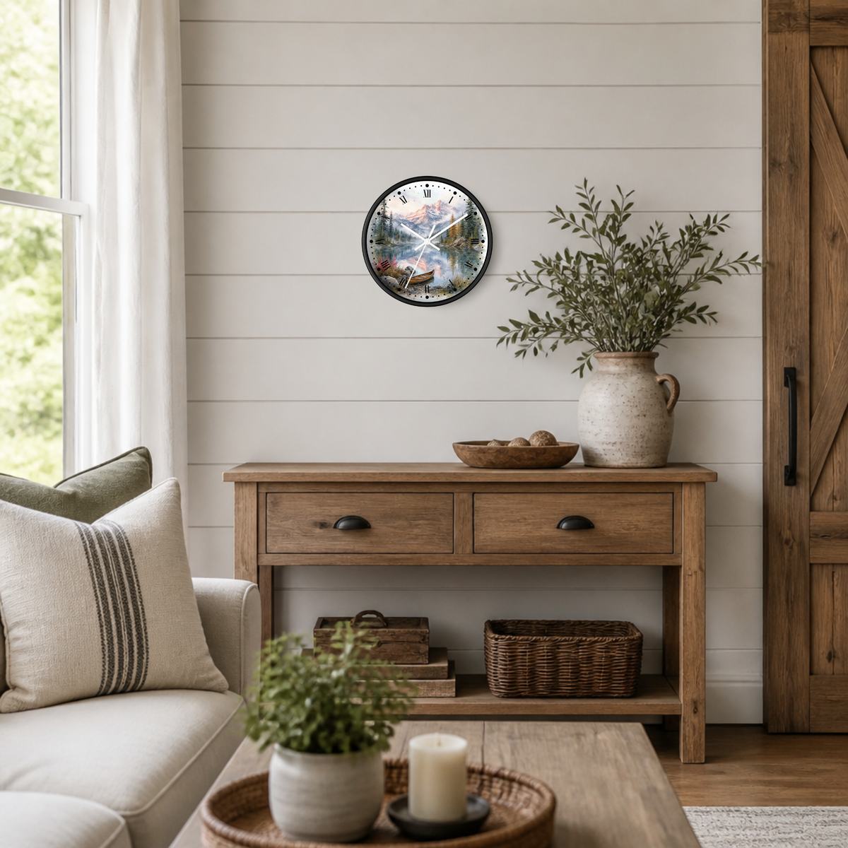 A round wall clock with a black frame and a detailed floral mountain lake illustration, styled on a white shiplap wall above a rustic wooden console table in a bright, modern farmhouse living room.
