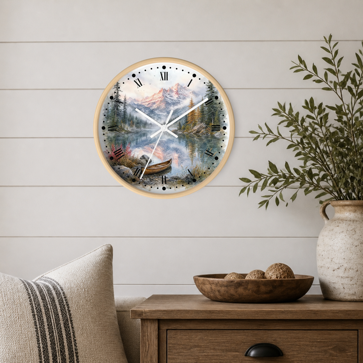 A round wall clock with a light wood frame and a scenic mountain lake print, mounted on a white shiplap wall above a wooden side table with a vase and bowl.