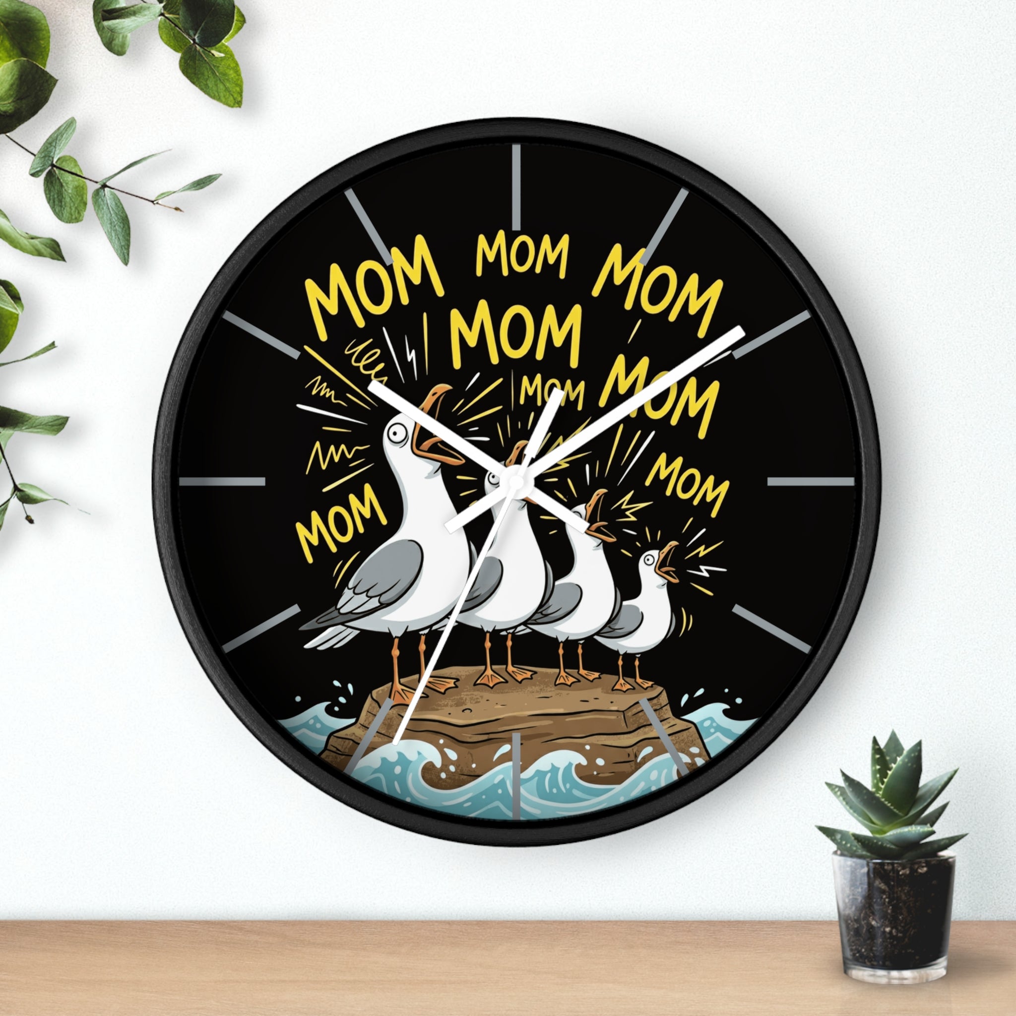 Round black wall clock with seagulls and 'Mom' text on a black background, placed on a surface with a plant.
