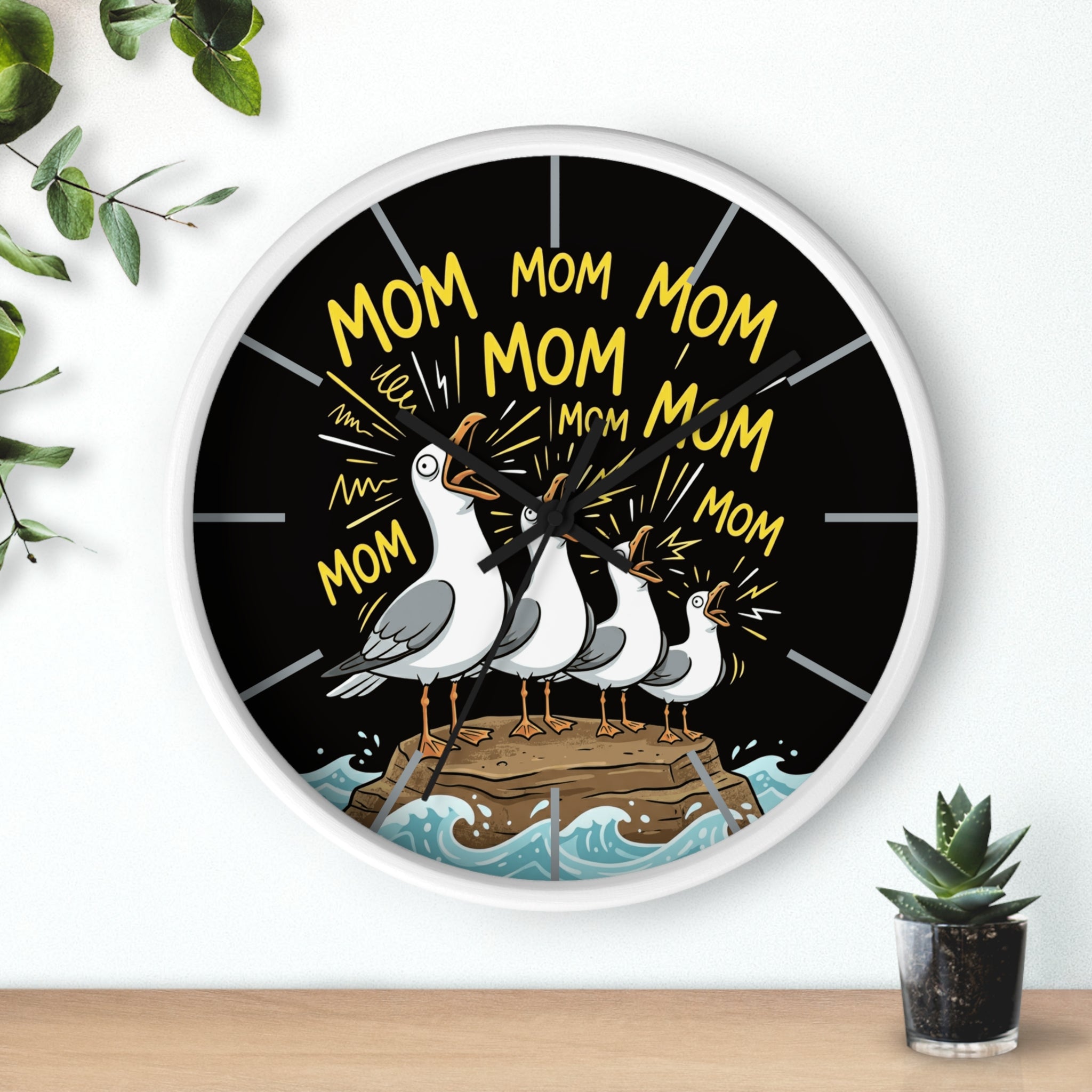 Round white wall clock with seagulls and 'Mom' text on a black background, placed on a white surface with a plant.