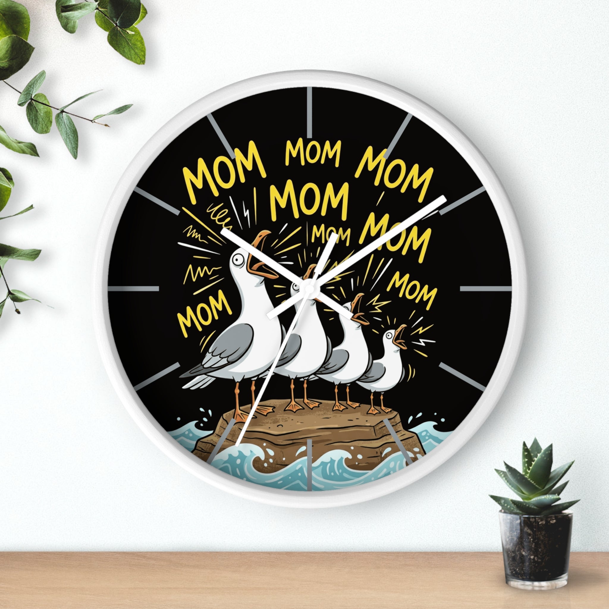 Round white wall clock with seagulls and 'Mom' text on a black background, placed on a white surface with a plant in the background.
