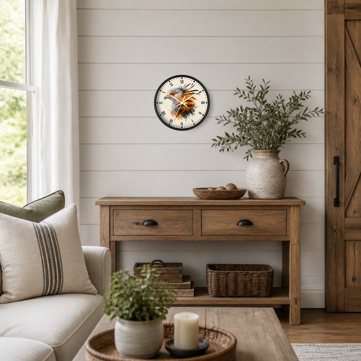 A round wall clock with a rustic illustration of an African secretary bird and cobra on the  face, styled on a white shiplap wall above a wooden console table in a bright, modern farmhouse living room.